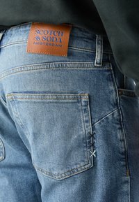 Blauwe denim jeans hebben een bruine leren patch met een reliëf logo van het merk, contrasterende stiknaad en een uniek kruissteeksysteem op de zak.