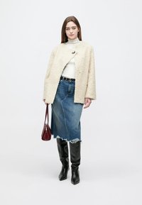Beige, pluizige oversized jas, witte gebreide coltrui, blauwe denim midi-rok met rafelige zoom, zwarte kniehoogte laarzen en een kleine bordeauxrode handtas.