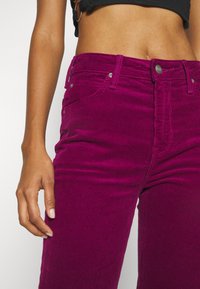 Femme portant un pantalon en velours côtelé magenta taille haute et un top court noir, la main gauche détendue le long du corps.