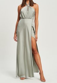 Frau in einem bodenlangen, hellgrauen Halskleid mit hohem Schlitz, der ihr linkes Bein zeigt, und beigen High Heels.