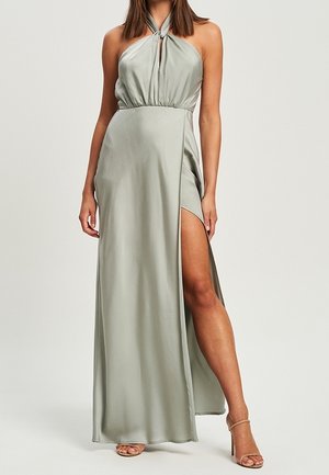 Femme portant une robe halter gris clair, longue jusqu'au sol, avec une fente haute révélant sa jambe gauche et des sandales à talons beiges.