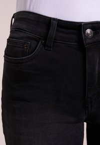 Jeans de denim negro con un diseño ajustado, bolsillos frontales visibles, herrajes de botón plateados y detalles de costura sutiles.