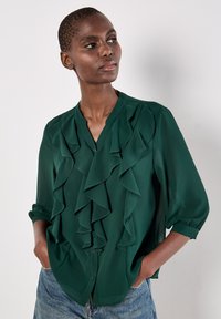 Blusa verde con dettagli arricciati, maniche lunghe arrotolate fino al gomito, chiusura a bottoni e tessuto liscio su uno sfondo chiaro.