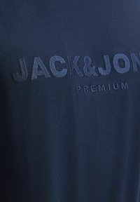 Tums zila kokvilnas T-kreklu ar izceltiem izšūtiem vārdiem "JACK&JON" un "PREMIUM" līdzīgās tumšās krāsās. Gluda tekstūra un vienkāršs dizains.
