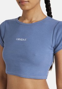 Top court côtelé bleu à manches courtes, avec un logo RVCA blanc sur le devant et une étiquette sur le côté. Texture lisse, coupe ajustée.