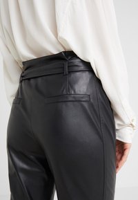Zwarte neplederen broek met hoge taille, voorzien van achterzakken en een gladde textuur, gecombineerd met een witte blouse.