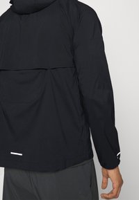 Mężczyzna w czarnej kurtce z długim rękawem, mającej wentylowane plecy i odblaskowe akcenty na mankietach oraz dolnej części pleców, w zestawieniu z ciemnymi spodniami.