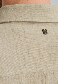 Lichtbeige shirt stof met een gestructureerde weving, voorzien van een zwart metalen logo op de achterkant van de kraag. Eenvoudig ontwerp, zonder patronen of accenten.