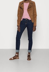 Chaqueta de motorista de ante marrón, camiseta rosa, jeans skinny oscuros y sandalias metálicas con tira en el tobillo. Texturas suaves y detalle de puños enrollados.
