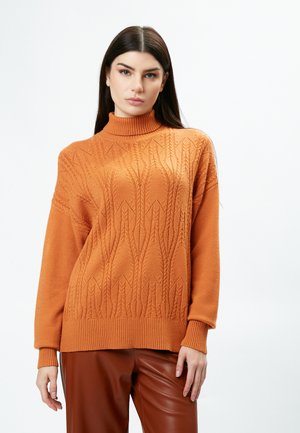 Gebreide coltrui in verbrand oranje. Heeft een textuurpatroon met een losse pasvorm en geribde manchetten en zoom. Gecombineerd met een paarse broek.