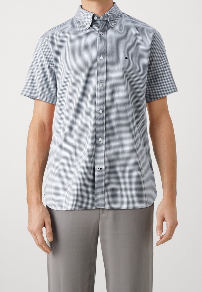 Homme portant une chemise à manches courtes à petits carreaux et un pantalon gris, debout devant un fond uni.