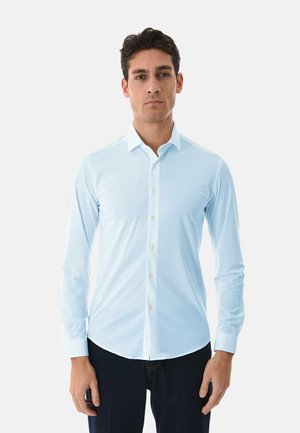 Uomo in piedi di fronte, indossa una camicia azzurra chiusa con bottoni a maniche lunghe e pantaloni scuri, sfondo uniforme.