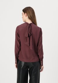 Maroon blouse with a bow detail at the back, long sleeves, button cuffs, and a smooth fabric. Worn with black leather pants.