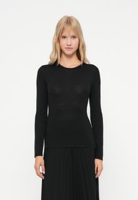 Γυναίκα με ξανθά μαλλιά, ντυμένη με εφαρμοστό μαύρο τοπ μακρυμάνικο και μαύρη πλισέ φούστα, που στέκεται μπροστά σε απλό λευκό φόντο.