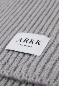 Tissu tricoté gris avec une texture côtelée ; présente une étiquette blanche avec un texte noir indiquant "ARKK COPENHAGEN." Perspective en gros plan.