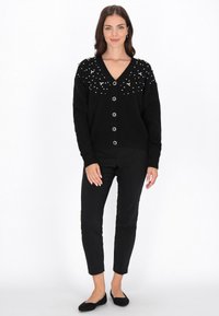 Czarny kardigan z dekoltem w kształcie litery V, ozdobiony rozrzuconymi dekoracyjnymi akcentami na ramionach, w zestawieniu z czarnymi dopasowanymi spodniami i płaskimi butami.