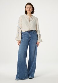 Crème blouse met blauwe en zwarte ster borduursels, knoopsluiting aan de voorkant en lange mouwen, gecombineerd met yüksek taille, wijde blauwe denim jeans.