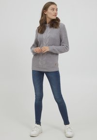 Szürke kötött pulóver texturált mintával és kábelmintával, kék skinny farmerral és fehér sportcipővel párosítva.