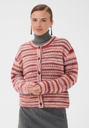 Jeune femme portant un cardigan tricoté rayé rose et rouge par-dessus un pull à col roulé gris et une jupe grise à fines rayures.