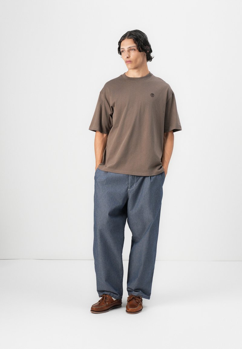 T-shirt en coton marron à manches courtes et col rond, assorti à un pantalon en denim bleu ample et des chaussures bateau marron.