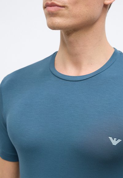 Άνδρας που φοράει μπλουζάκι σε πράσινο-γαλάζιο απόχρωση με στρογγυλή λαιμόκοψη και λευκό λογότυπο αετού στο αριστερό στήθος, φαίνεται από τους ώμους μέχρι το πηγούνι με φόντο λευκό.