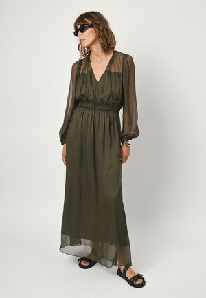 Olivengroene maxi-jurk met doorschijnende lange mouwen, een getailleerde taille en een diepe V-hals. Lichte stof valt soepel en is gecombineerd met zwarte sandalen.