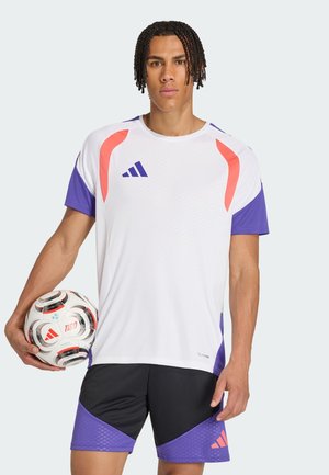 Mandlig atlet i hvidt og lilla sportstøj holder en fodbold og står mod en ensfarvet lys baggrund.