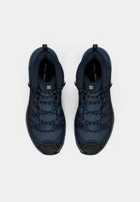 Et par med marineblå og sorte tursko med snørebånd, polstrede krager og logo på tunga sett ovenfra på en hvit bakgrunn.