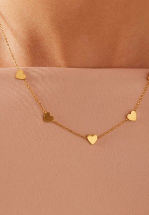 Gouden ketting met gelijkmatig verdeelde kleine hartvormige hangers, gedragen op een sleutelbeen boven een beige strapless top.