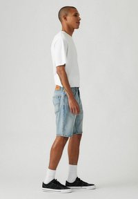 Weißes T-Shirt mit kurzen Ärmeln, hellblaue Jeansshorts und schwarze Turnschuhe mit weißen Sohlen. Die Shorts haben eine lockere Passform und leicht ausgefranste Saumkanten.