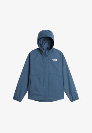 Meisje draagt een blauwe jas, zwarte legging, witte muts, zonnebril, witte sokken, wandelschoenen en draagt een zwarte rugzak.