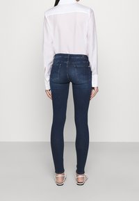 Jeans ajustados de color azul oscuro con una cintura alta, cinco bolsillos y detalles de costura sutiles, combinados con una camisa ligera de manga larga blanca.