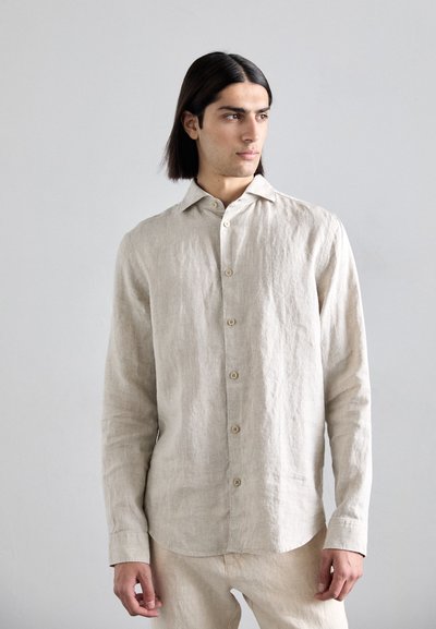 Chemise en lin beige avec un devant à boutons, des manches longues, un col pointu et un ourlet légèrement incurvé. Tissu texturé simple et couleur neutre.