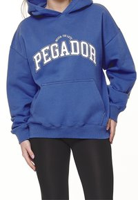 Osoba oblečená v modré mikině s nápisem "Nikdy není pozdě PEGADOR" a černých legínách, ruce uvolněně po stranách.