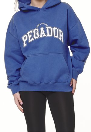Persona vestida con una sudadera azul que dice "Never Too Late PEGADOR" y leggings negros, con las manos relajadas a los lados.
