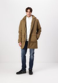 Khaki parka jas met een capuchon, voorste drukknoopzakken en een binnenvoering, gecombineerd met een witte fleece met rits en donkere jeans. Zwarte sneakers.