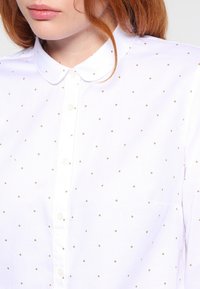 Camisa blanca de botones con collar puntiagudo y patrones de diamantes dorados. La tela tiene un acabado texturizado y un bolsillo en el pecho.