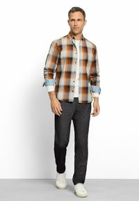 Mann läuft und trägt ein orange-blaues kariertes Hemd über einem weißen T-Shirt, dunkle graue Hosen und weiße Turnschuhe vor einem weißen Hintergrund.