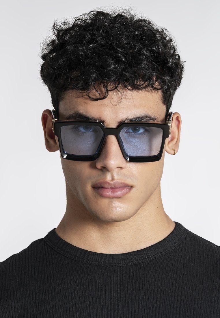 Junger Mann mit lockigem Haar trägt große, quadratische schwarze Sonnenbrillen und ein schwarzes, geripptes Hemd, blickt nach vorne vor einem schlichten, hellen Hintergrund.