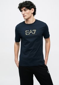 Tengerészkék pamut póló arany logóval és csíkokkal, kerek nyakkivágással és rövid ujjakkal, fekete nadrággal párosítva.