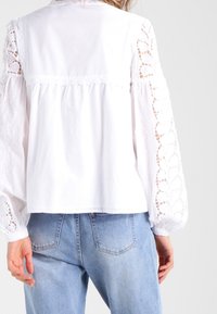 Blouse blanche avec des manches longues en dentelle brodée, coupe décontractée et design froncé au dos, associée à un jean en denim bleu clair.