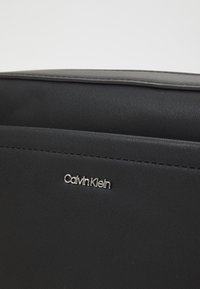 Sac noir Calvin Klein avec emblème logo argenté centré sur un matériau texturé en gros plan.