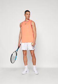 Giocatore di tennis maschile che tiene una racchetta e una palla, indossando una maglietta senza maniche arancione, pantaloni bianchi, calze e sneakers bianche Nike su uno sfondo neutro.
