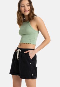 Světlý zelený žebrovaný crop top se zvlněným lemem, v kombinaci s černými šortkami na šňůrku, které mají knoflíky po stranách a boční kapsy.