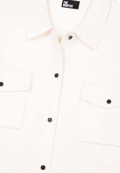 Weißes Hemd aus strukturiertem Stoff mit Kragen, zwei Vorderentaschen und schwarzen Blütenverzierungen als Knöpfe.