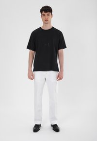 Black cotton t-shirt with short sleeves and a round neckline, paired with white trousers and black footwear. Simple design, no patterns.