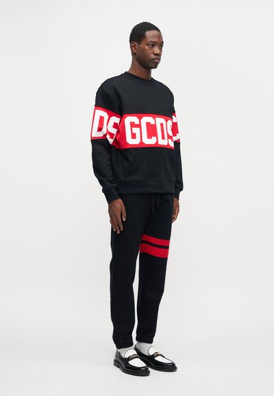 Μαύρη φούτερ με μεγάλο άσπρο και κόκκινο γράμμα, συνδυασμένη με μαύρο παντελόνι με κόκκινες ρίγες. Το μοντέλο φ wears γυαλιστερά μαύρα loafers.