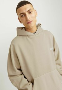 Béžová oversized mikina s kapucí, přední kapsou, měkkou texturou a volným střihem. Na hrudi se nachází malé logo.