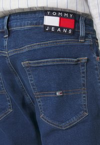 Dark blue denim jeans featuring a logo patch on the back waistband with red, white, and blue accents, and standard back pockets with stitching details.
