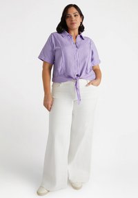 Camicia button-up a maniche corte di un leggero colore viola, con un nodo in vita, abbinata a pantaloni bianchi a gamba larga e scarpe beige, caratterizzata da una texture liscia.